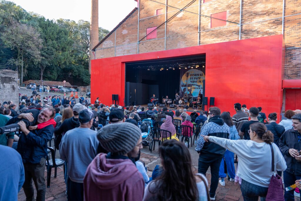 Foto em ângulo aberto mostra o público do festival, com pessoas sentadas e em pé em frente ao Teatro do Engenho, de fachada vermelha. O palco ao fundo exibe músicos tocando, enquanto a plateia lota o espaço externo, criando clima de confraternização. Algumas pessoas aparecem conversando e sorrindo, em ambiente descontraído.