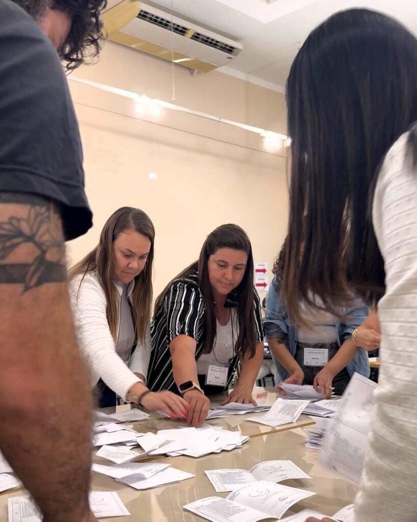 Um close mostra pessoas contando e organizando as cédulas de votação em cima de uma mesa. Duas mulheres, uma de cabelo loiro e outra de cabelo castanho, se concentram nos papéis, enquanto outras pessoas ao redor ajudam no processo.
