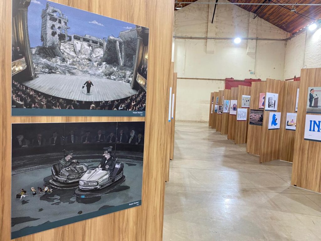 Painéis exibindo cartuns de artistas estrangeiros. Um mostra um homem de cartola em meio a destroços, diante de uma plateia aplaudindo. Outro apresenta dois políticos em carrinhos de bate-bate, observados por uma plateia. O espaço é amplo, de pé-direito alto e paredes claras.