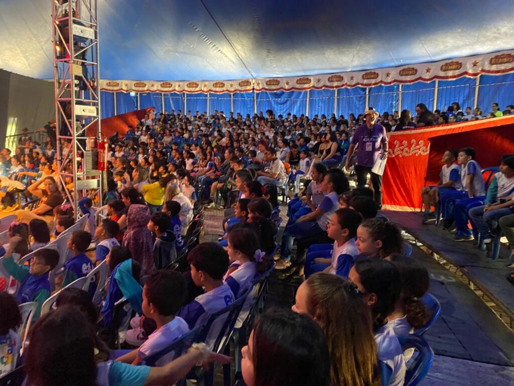Vista ampla de dentro da lona do circo, mostrando centenas de crianças uniformizadas ocupando todas as arquibancadas. A cena transmite energia, diversidade e a grandiosidade do evento, com o espaço totalmente tomado pelo público.