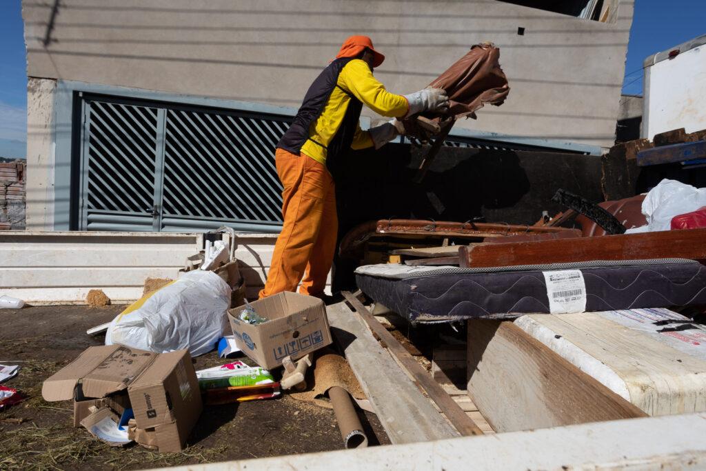 Um trabalhador com roupas de proteção, incluindo calças laranjas e um boné, está descartando móveis em um caminhão de lixo. Ele está segurando um pedaço de couro marrom, possivelmente de um sofá ou cadeira, e o está jogando no caminhão. O caminhão está cheio de outros itens de descarte, como colchões, pedaços de madeira e sacos de lixo. Há também caixas de papelão e outros detritos espalhados no chão ao redor do caminhão. O cenário parece ser uma área urbana, com uma casa ou prédio ao fundo e um céu azul claro acima.