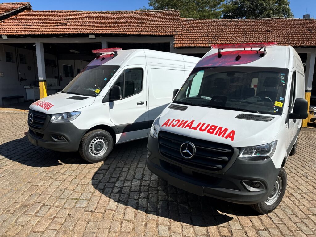 A imagem mostra duas ambulâncias brancas da Mercedes-Benz estacionadas sobre um piso de paralelepípedos, em frente a um prédio com telhado de cerâmica. Ambas têm a palavra “AMBULANCIA” escrita em vermelho na parte frontal, destacando sua função de atendimento emergencial. Ao fundo, o edifício parece ser uma garagem ou área de serviço, com baias abertas e alguns equipamentos visíveis no interior.
