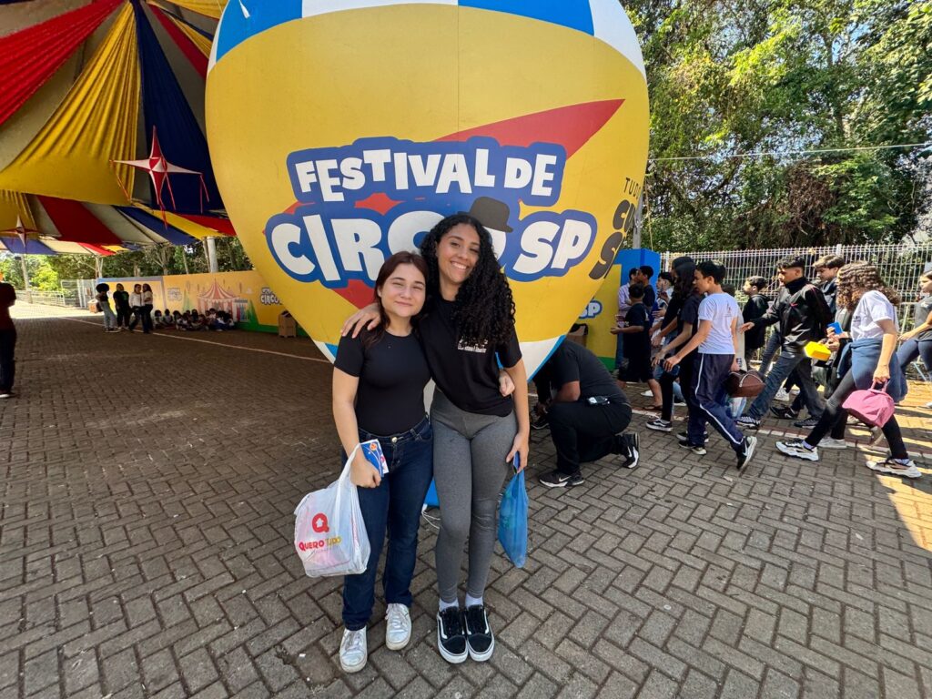 Pra cego ver: Duas adolescentes posam sorridentes abraçadas em frente a um balão cenográfico amarelo e azul com o logotipo “Festival de Circo SP”. Atrás delas, outros estudantes caminham pelo espaço do evento.