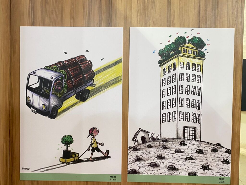 À esquerda: um caminhão pequeno transporta toras de madeira. Atrás dele, uma pessoa puxa um carrinho de mão com um vaso de planta, sugerindo contraste entre desmatamento e preservação. À direita: um prédio alto, branco, cheio de janelas. No topo, há árvores e pássaros, como se fosse uma floresta suspensa. Abaixo, o chão rachado e cheio de tocos de árvores cortadas, reforçando a crítica ambiental.