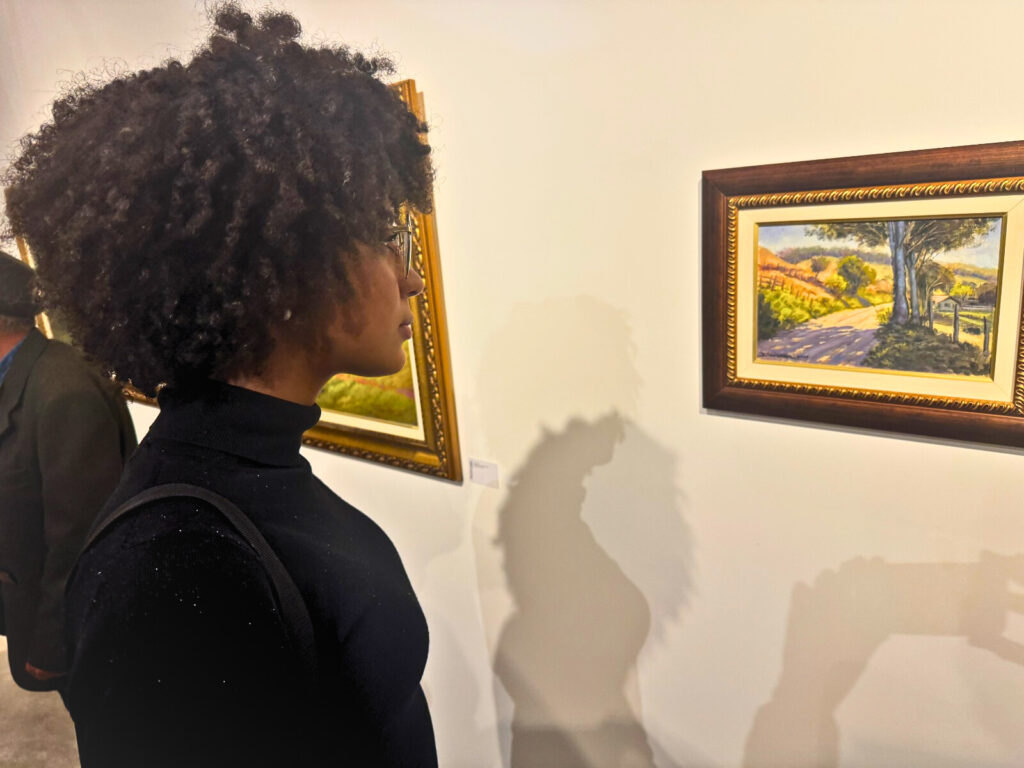 Mulher de cabelos cacheados observa de perto um quadro com paisagem de estrada e árvores.
