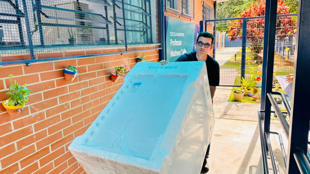 A imagem mostra uma pessoa carregando uma máquina de lavar roupas branca, em uma área externa de uma escola municipal chamada "Professor Matheus Tullio". O ambiente é cercado por um muro de tijolos com pequenos vasos de plantas pendurados, além de outras plantas ao fundo. Há uma grade metálica com um portão que dá acesso a outra parte do terreno escolar.