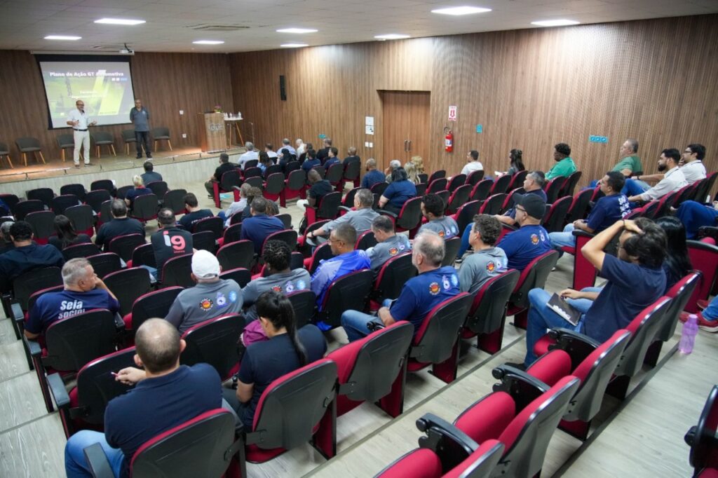 A imagem mostra uma sala de conferências com uma audiência sentada em cadeiras vermelhas e pretas, voltada para um palco. No palco, há dois homens em pé, um deles falando para a audiência. Há também uma tela de projeção com texto em português, que parece ser um plano de ação. A sala tem paredes de madeira e iluminação no teto. A audiência é composta por homens e mulheres de diversas idades.