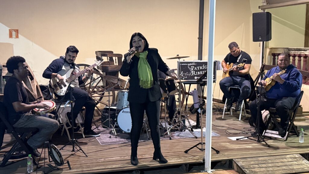Banda com cinco músicos se apresenta; uma cantora de cachecol verde canta ao centro, acompanhada por músicos tocando instrumentos como violão, cavaquinho, contrabaixo e pandeiro.
