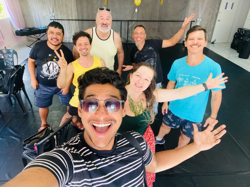 A imagem mostra um grupo de oito pessoas em uma sala com piso escuro e paredes claras. A maioria das pessoas está sorrindo e olhando para a câmera. Na frente, um homem com óculos escuros e uma camiseta listrada preta e branca está tirando uma selfie. Ele tem um sorriso largo e está acenando com a mão. Atrás dele, há mais sete pessoas. Da esquerda para a direita, um homem em uma camiseta preta com um desenho branco, um homem em uma camiseta amarela, um homem mais velho em uma regata bege, um homem em uma camiseta preta, uma mulher em uma regata verde com um colar verde, e um homem em uma camiseta azul. Todos parecem estar se divertindo. No fundo, há uma barra de dança, um teclado e algumas cadeiras.