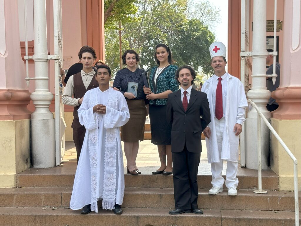 Um grupo de seis pessoas está em pé em uma escada em frente a um edifício com paredes rosas e colunas brancas.Da esquerda para a direita, as pessoas são:* Uma pessoa com um colete marrom sobre uma camisa clara.* Uma pessoa vestindo uma túnica religiosa branca com uma estola branca adornada com padrões intrincados e franjas, segurando um rosário.* Uma mulher com uma blusa escura estampada e uma saia marrom.* Uma mulher com uma saia escura e uma jaqueta com padrões em teal e preto. Ela está segurando dois livros.* Um homem em um terno preto com uma gravata vermelha.* Uma pessoa vestida de médico, usando um jaleco branco, calças brancas, sapatos brancos e um gorro branco com uma cruz vermelha. Também está usando uma gravata vermelha.As pessoas parecem estar posando para uma fotografia. A iluminação sugere que é de dia.