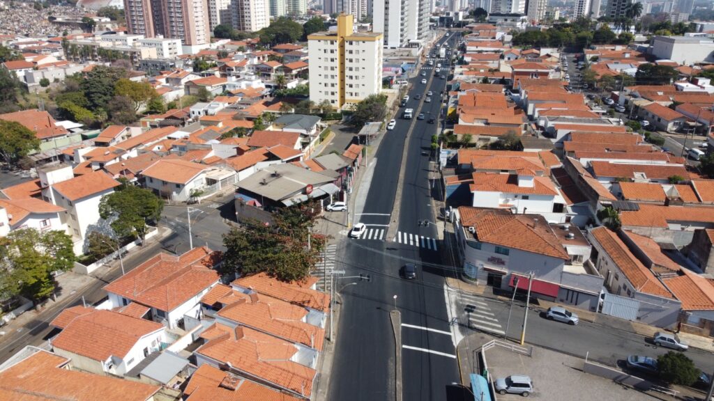 A imagem apresenta uma vista aérea de uma área residencial com casas de telhados vermelhos organizadas em quadras bem definidas. Uma avenida principal corta o centro da cena, com faixas de pedestres e sinais de trânsito visíveis, indicando boa infraestrutura urbana. Alguns carros circulam pela via, sugerindo movimento moderado. Ao fundo, surgem prédios mais altos, contrastando com a paisagem predominantemente horizontal das casas, o que revela uma transição entre zona residencial e área mais urbanizada.