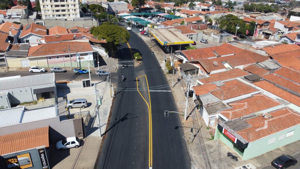 A imagem mostra uma vista aérea de um bairro urbano com ruas bem definidas e casas de telhados vermelhos. A via principal está recém-pintada com faixas amarelas, indicando organização no tráfego. À direita, destaca-se um posto de gasolina da marca Ipiranga, com cobertura amarela e estrutura visível. Há alguns carros estacionados e movimentação discreta, sugerindo um ambiente tranquilo e funcional.