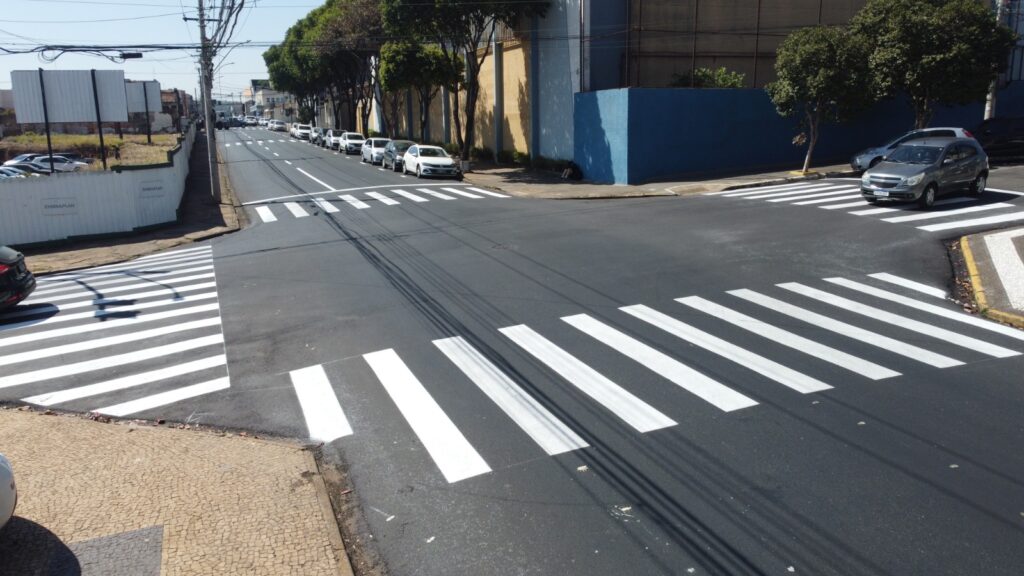 A imagem mostra um cruzamento urbano com faixas de pedestres recém-pintadas em branco, destacando o cuidado com a sinalização e a segurança viária. Há carros estacionados ao longo da rua e construções ao redor, incluindo uma cerca branca com o nome “EMBRAPLAN”, sugerindo a presença de uma empresa ou obra local. Árvores e postes com fiação elétrica compõem o cenário, reforçando o ambiente de cidade.