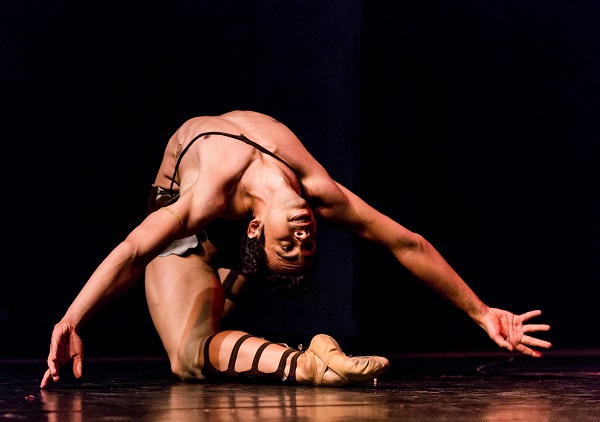 Pra Cego Ver: O bailarino Alan Marques está sozinho no palco, em um movimento expressivo de arco para trás. Ele veste figurino de dança em tom bege, com tiras que lembram sandálias gladiadoras. Seu corpo está arqueado, uma mão toca o chão e a outra se estende para trás, enquanto seu rosto olha em direção ao público, transmitindo intensidade e emoção.
