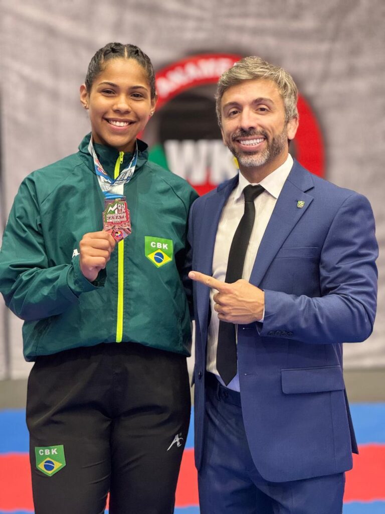 Na imagem, uma jovem atleta sorri orgulhosamente enquanto segura uma medalha. Ela veste um agasalho verde com detalhes amarelos e calças pretas, ambos adornados com o emblema da Confederação Brasileira de Karatê (CBK). Ao lado dela, um homem de terno azul, com uma gravata preta, aponta para a medalha, compartilhando o momento de celebração. O fundo sugere um ambiente de competição, possivelmente um campeonato de karatê, com um logotipo circular em vermelho e branco ao fundo.