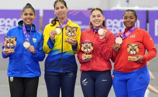 A imagem mostra quatro mulheres em um pódio, segurando medalhas e troféus. Da esquerda para a direita, a primeira mulher, com a pele mais clara, veste um agasalho azul com detalhes verdes e pretos, ostenta uma medalha de prata e um troféu com a figura de um gato. A segunda mulher, com a pele mais clara, veste um agasalho amarelo com detalhes azuis e pretos, exibindo uma medalha de ouro e um troféu semelhante. A terceira mulher, com a pele mais clara, usa um agasalho vermelho e calças de compressão azuis escuras, segurando uma medalha de bronze e um troféu de gato. A quarta mulher, com a pele mais escura, veste um agasalho vermelho com a escrita "USA" e calças de agasalho azuis, também segurando uma medalha de bronze e um troféu de gato. Todas as mulheres sorriem para a câmera, demonstrando satisfação com suas conquistas. O pódio é roxo e há letras desfocadas ao fundo.