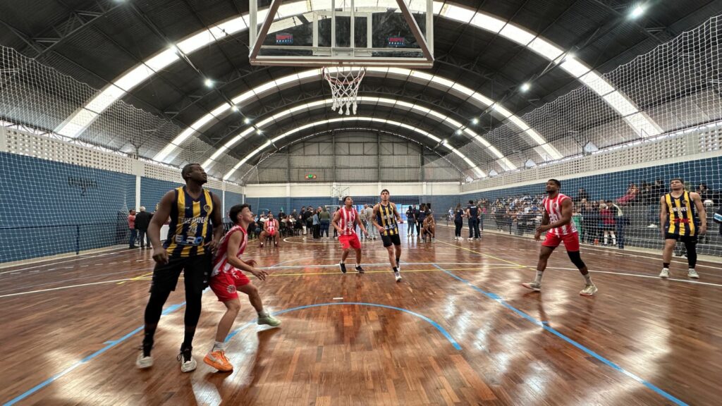 A imagem mostra uma partida de basquete em uma quadra interna. Vários jogadores estão em ação, vestindo uniformes de duas cores distintas: um time usa uniformes vermelhos com listras brancas, enquanto o outro usa uniformes amarelos com listras pretas. A quadra possui um piso de madeira brilhante e as linhas de jogo são visíveis.O teto da quadra é arqueado e possui iluminação artificial, com algumas janelas no topo permitindo a entrada de luz natural. Uma rede de proteção está instalada ao redor da quadra, acima das arquibancadas onde espectadores estão sentados, assistindo ao jogo. A atmosfera parece ser de um evento esportivo animado.