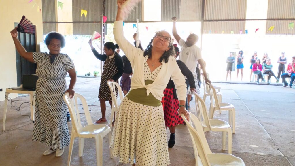 A imagem mostra uma atividade coletiva em um espaço interno, onde várias pessoas — em pé, com os braços erguidos e segurando leques — participam de um momento interativo ou festivo. O local é amplo, com cadeiras plásticas organizadas e bandeirinhas coloridas penduradas no teto, reforçando o clima de celebração.