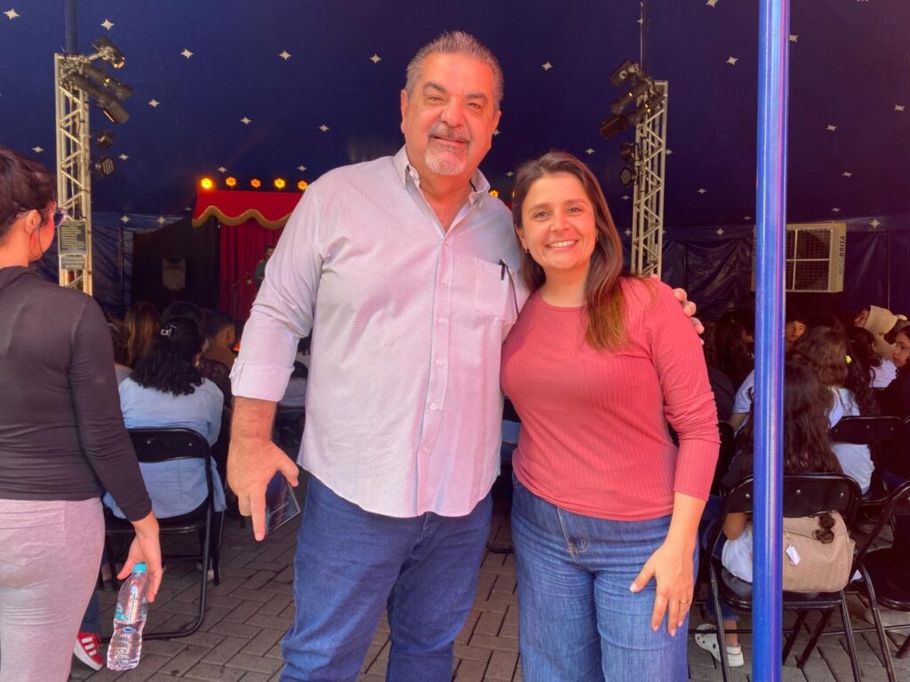 O secretário de Cultura, Carlos Beltrame, e a secretária de Turismo, Clarissa Quiararia, posam sorridentes para a câmera em frente à plateia do circo. Beltrame veste camisa social clara e Clarissa usa blusa vermelha de manga comprida.