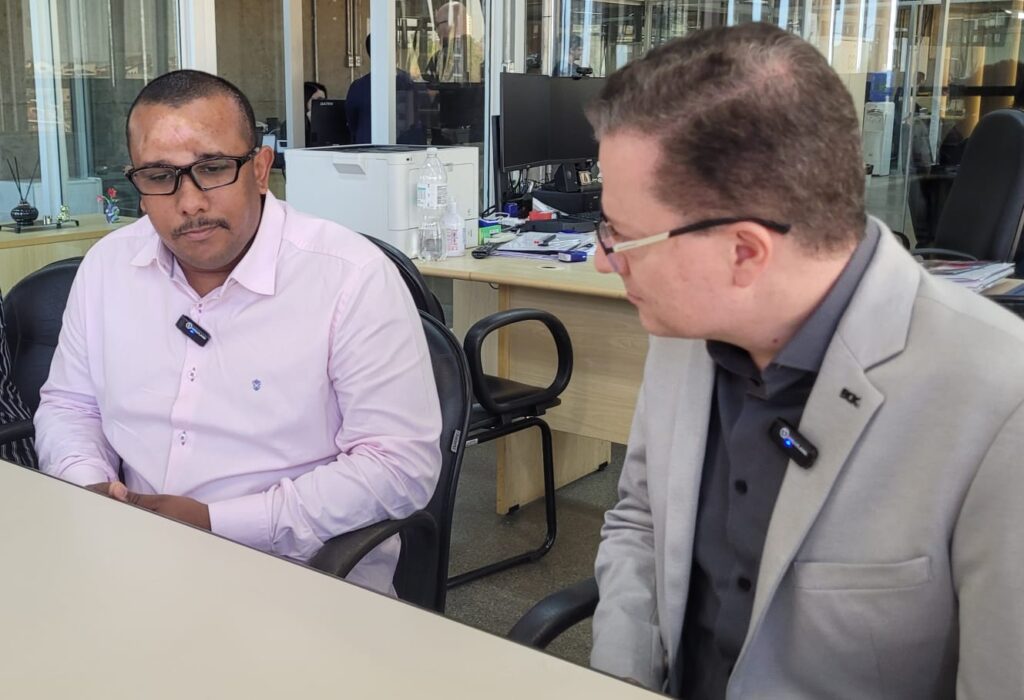 Dois homens estão sentados em uma sala, com uma mesa à frente deles.À esquerda, um homem com pele escura e óculos escuros está vestindo uma camisa rosa clara. Ele tem um microfone preso à sua camisa.À direita, um homem com pele clara e óculos está vestindo um terno cinza claro sobre uma camisa cinza escura. Ele também tem um microfone preso à sua lapela.Ao fundo, há uma mesa com um computador, uma impressora e uma garrafa de água. Há também outras pessoas e escritórios visíveis através de uma divisória de vidro.