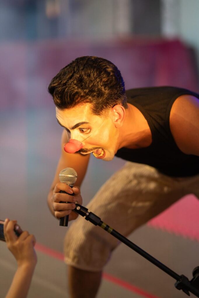 Homem maquiado como palhaço com nariz vermelho se inclina em direção a um microfone, com expressão intensa e teatral. Ele segura o microfone e canta próximo a alguém que o filma com um celular. A iluminação cênica destaca seu rosto e transmite energia e emoção da performance.