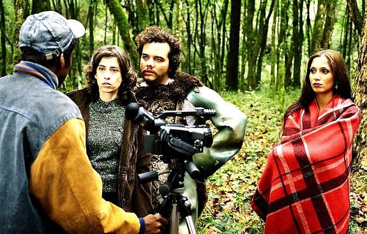 Quatro pessoas em uma floresta conversam próximas a uma câmera; uma mulher usa manta xadrez vermelha, outra encara o interlocutor com expressão séria, e um homem com casaco peludo observa.