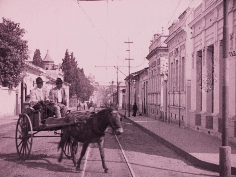 Fotografia em tom sépia mostra uma rua antiga de Piracicaba no início do século XX. Dois homens, vestidos com ternos claros e chapéus, estão sentados em uma charrete puxada por um cavalo que avança em direção à câmera. A via é ladeada por casas e prédios de arquitetura colonial, com postes e fios de bonde elétrico no centro da rua. A cena transmite a atmosfera da cidade em 1922.