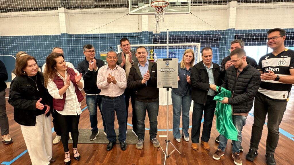 A imagem mostra um grupo de pessoas em um ginásio esportivo, possivelmente durante uma inauguração ou evento. Há uma placa com texto em destaque no centro, com um homem de camisa azul e jaqueta escura apresentando-a, enquanto outros ao redor aplaudem. Uma pessoa a sua direita, com uma camisa preta e branca com o número "XV" e a palavra "raízen", também aplaude. Ao fundo, pode-se ver uma quadra de basquete com uma rede e uma cesta. O ambiente parece ser um evento oficial, com a presença de autoridades e convidados.