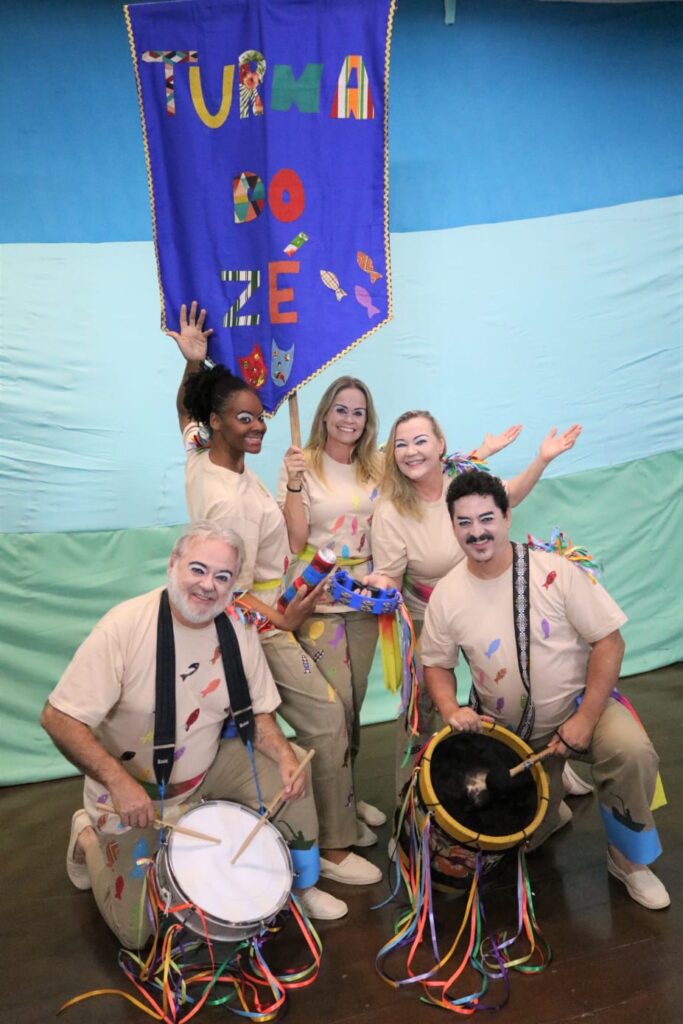 Cinco integrantes do grupo posam sorrindo em frente a um fundo azul, segurando uma bandeira com letras coloridas que formam o nome “Turma do Zé”. Todos usam figurino com desenhos coloridos, maquiagem teatral e acessórios com fitas coloridas. Dois deles estão com instrumentos de percussão, como tambor e caixa. O grupo transmite alegria e energia típica do teatro popular.