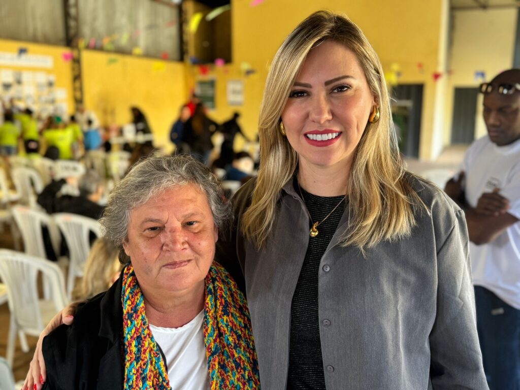 Claro! A imagem destaca duas pessoas em pé, lado a lado, em um ambiente interno decorado com enfeites coloridos e cheio de cadeiras brancas. A pessoa à esquerda usa um casaco escuro e um lenço colorido vibrante, que chama atenção, transmitindo um ar acolhedor e expressivo. Já a pessoa à direita veste uma camiseta cinza simples com um colar dourado, sugerindo um estilo mais discreto.