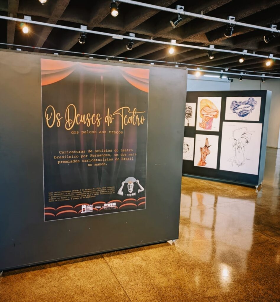 Foto da exposição “Os Deuses do Teatro” no hall do Teatro Municipal Losso Netto. Em destaque, um painel central preto com o cartaz da mostra, que traz o título em letras douradas e o subtítulo explicando: “Caricaturas de artistas do teatro brasileiro por Fernandes, um dos mais premiados caricaturistas do Brasil no mundo”. Ao redor, vê-se caricaturas emolduradas nas laterais, expostas em painéis pretos.