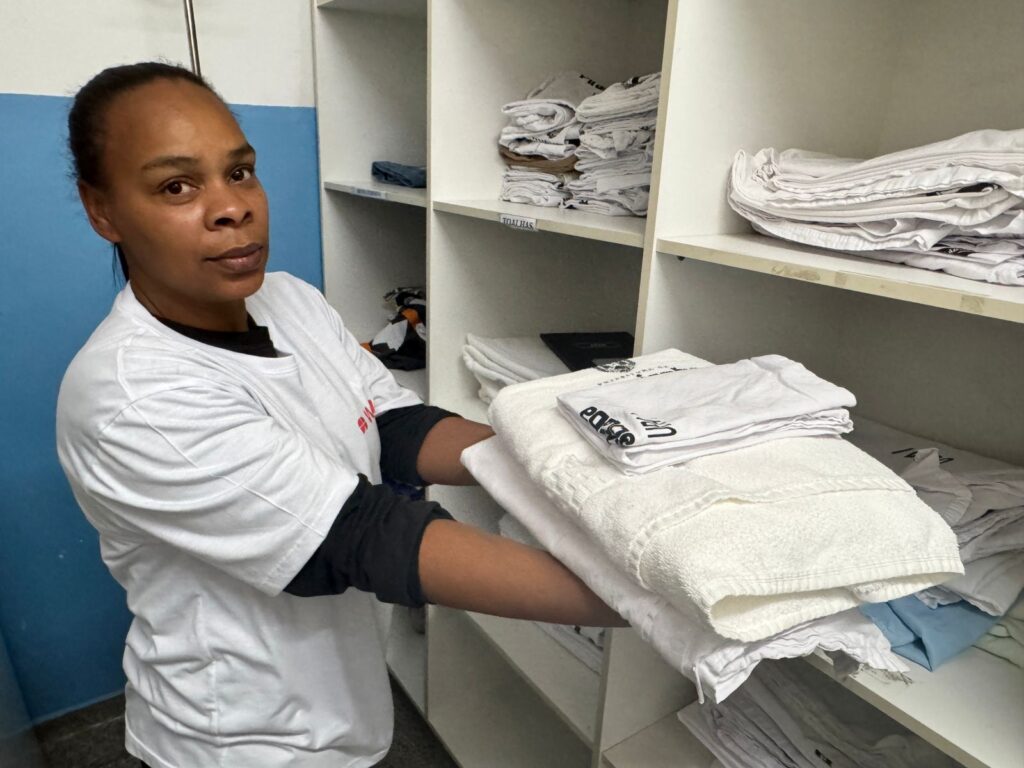 A mulher, com pele morena e cabelos presos para trás, usa uma camiseta branca com mangas compridas pretas por baixo. Ela segura uma pilha de toalhas brancas e uma camiseta branca com a palavra "eapge" escrita em letras pretas. Atrás dela, há prateleiras cheias de roupas brancas dobradas, incluindo o que parecem ser toalhas e camisetas. Uma prateleira mais baixa tem um par de jeans escuros e um pequeno objeto com o que parece ser um pinguim. A parede atrás dela é azul e branca.