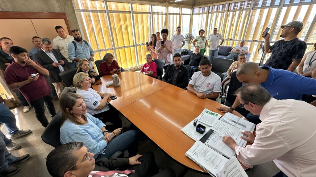 Sala cheia com pessoas sentadas e em pé ao redor de uma mesa de reuniões. No centro da mesa estão papéis e pastas de documentos. Algumas pessoas registram o momento com celulares enquanto acompanham a assinatura dos termos.