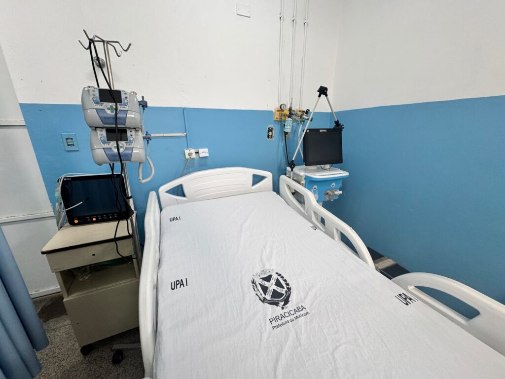 A imagem mostra um quarto de hospital com duas camas de hospital brancas e totalmente equipadas. As camas estão posicionadas lado a lado, com cortinas azuis separando-as e criando privacidade para cada paciente. Cada cama é de metal branco e possui grades laterais que podem ser levantadas para evitar que os pacientes caiam. O colchão de cada cama está coberto com um lençol branco, e ambos os lençóis parecem ter o logotipo de uma instituição ("UPA") impresso.