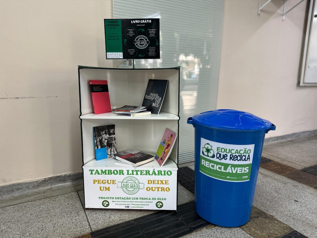 A imagem retrata um espaço dedicado à promoção da leitura e da sustentabilidade, com destaque para um tambor literário e uma lixeira de recicláveis. O tambor literário funciona como um ponto de troca de livros, incentivando o acesso gratuito à leitura por meio do lema “Pegue um, deixe outro”. Ele está repleto de livros e traz uma placa informativa com detalhes do projeto e contatos para doações. Ao lado, uma lixeira azul com os dizeres “Educação que recicla” e “Recicláveis” reforça o compromisso ambiental do local.