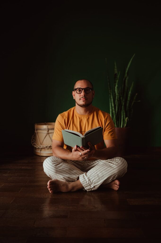Homem careca, de óculos, barba rala e pele clara, está sentado no chão de pernas cruzadas. Ele veste camiseta amarela e calça clara listrada, segura um livro aberto nas mãos e olha diretamente para a câmera. Atrás dele há uma parede verde-escura, um vaso com planta alta e um tambor de madeira.