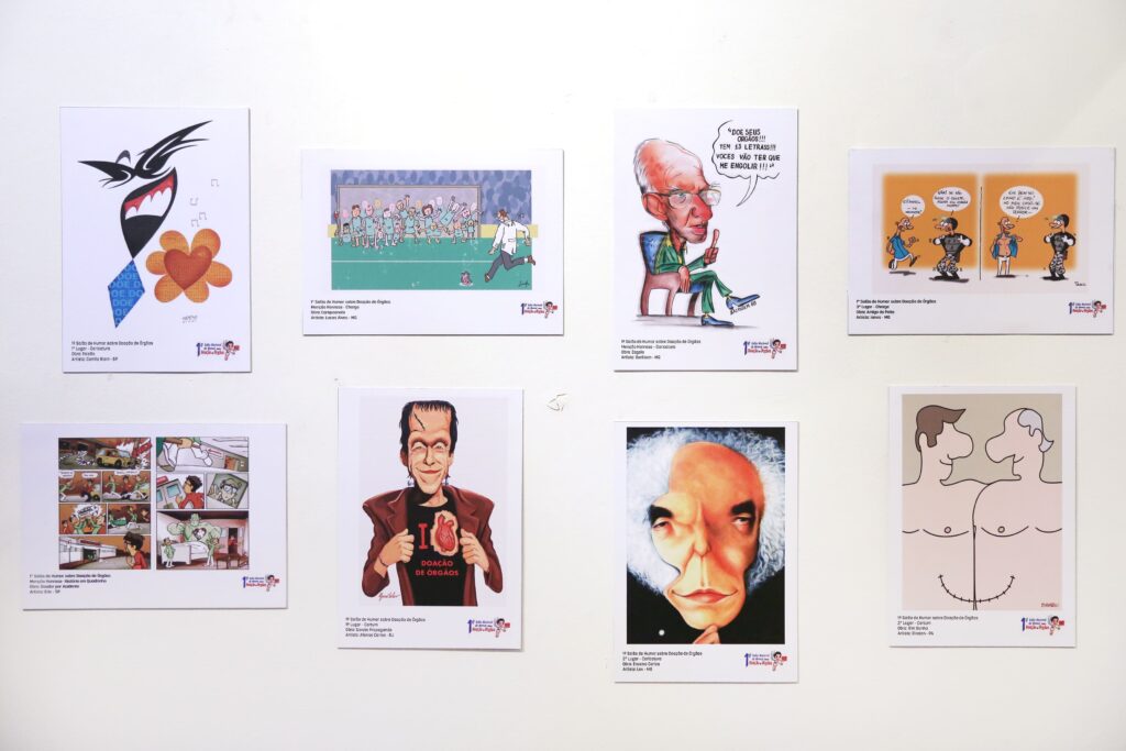 Descrição: Outra parede branca reúne oito cartuns e caricaturas coloridas sobre Doação de Órgãos. Entre eles, há desenhos de médicos, caricaturas de personalidades, uma equipe de futebol, uma figura segurando um coração e ilustrações estilizadas que destacam o tema da doação.