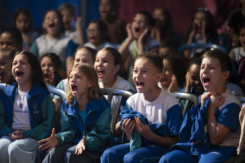 Grupo de crianças com uniforme escolar reage entusiasmado dentro da lona. Elas sorriem e gritam de empolgação com olhos arregalados.