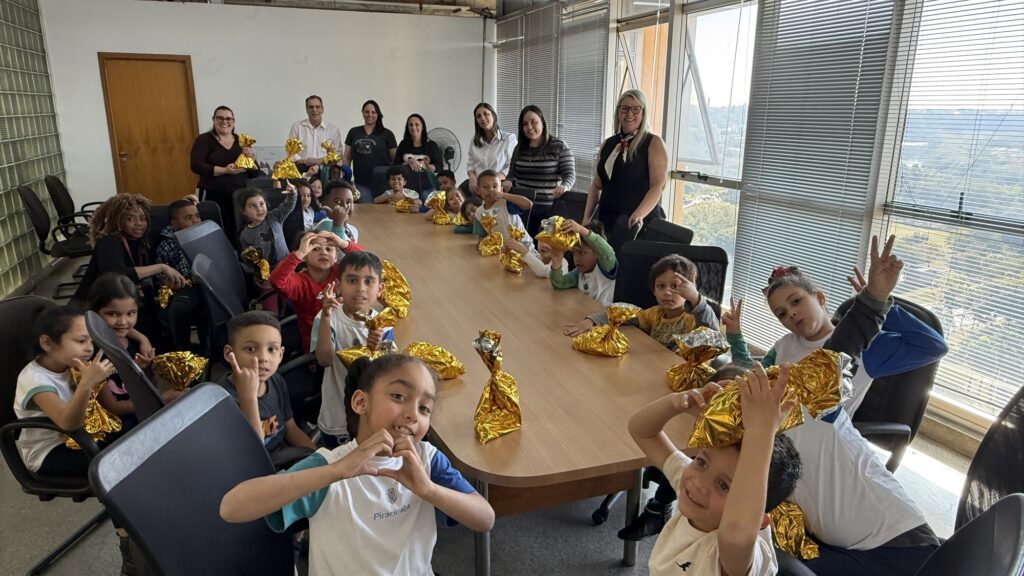 A imagem retrata um momento alegre e coletivo em uma sala de reuniões iluminada por luz natural, onde crianças e adultos estão reunidos ao redor de uma mesa grande. As crianças seguram pacotes embrulhados em papel dourado, sugerindo que estão recebendo presentes, talvez como parte de uma celebração ou evento especial. O ambiente é acolhedor, com janelas amplas, persianas, uma porta de madeira e uma parede de blocos de vidro que compõem o cenário.