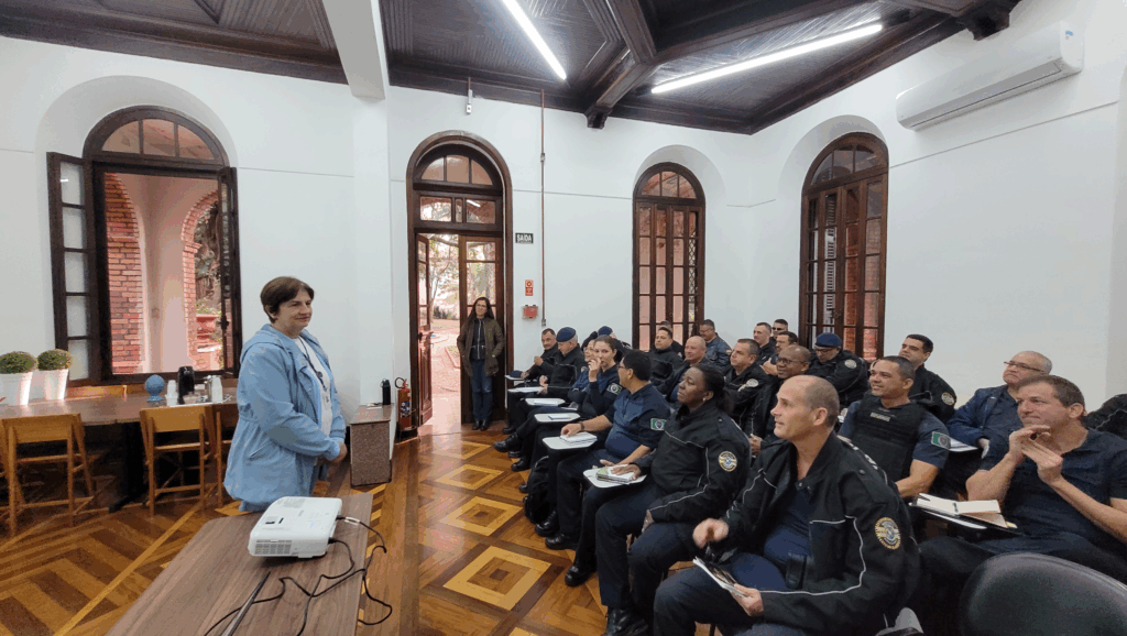 A imagem mostra uma sala de aula com um grupo de pessoas, a maioria vestindo uniformes pretos com emblemas amarelos, possivelmente policiais. Há uma mulher de pé à esquerda, vestida com um casaco azul claro, que parece estar apresentando algo. Ela está ao lado de uma mesa com um projetor.A sala tem um teto de madeira escura com iluminação embutida, paredes brancas e janelas altas e arqueadas com detalhes em madeira escura. Há também uma porta aberta à esquerda, que dá para uma área externa com tijolos aparentes e plantas.No lado direito da sala, há um ar condicionado instalado na parede. As pessoas estão sentadas em cadeiras, a maioria com cadernos ou papéis à sua frente, indicando que estão em uma aula ou treinamento. O piso é de madeira com um padrão geométrico em tons de marrom.