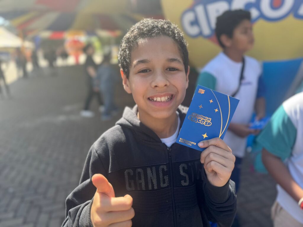 Pra cego ver: Um garoto de cerca de 10 anos sorri para a câmera, mostrando o passaporte azul do Festival de Circo. Ele veste moletom escuro com capuz e faz sinal de positivo com a outra mão. Ao fundo, outros colegas caminham pelo espaço.