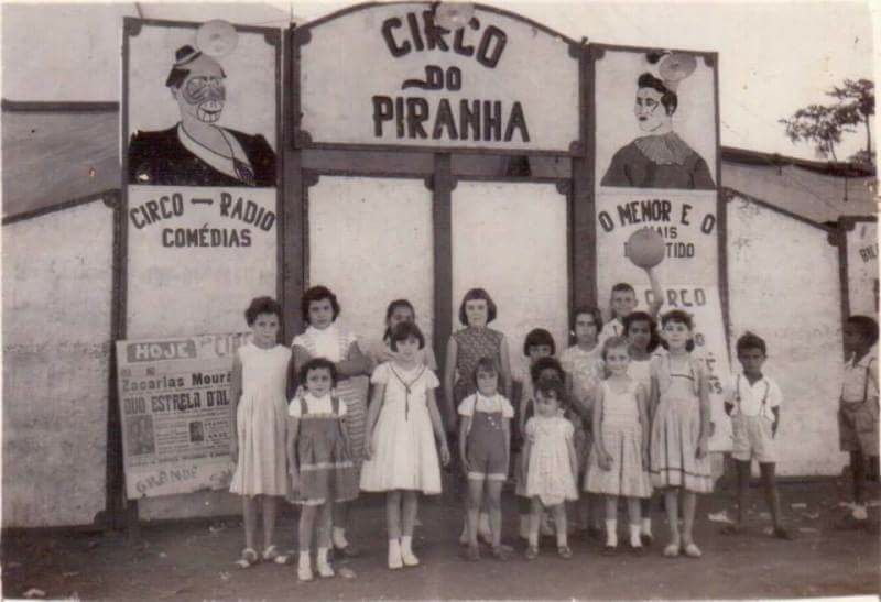 Foto antiga em preto e branco mostra grupo de crianças posando em frente à entrada do “Circo do Piranha”, com cartazes ilustrados nas laterais.