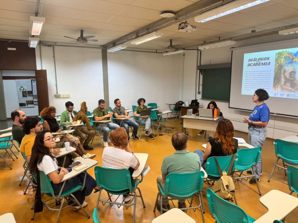 A imagem retrata um ambiente acadêmico ou profissional, onde um grupo de pessoas está reunido em formato de semi-círculo, participando de uma apresentação intitulada "DIÁLOGOS COM A ACADEMIA". O palestrante está em pé diante dos participantes, com um slide projetado que exibe a imagem de um macaco e diversos logotipos institucionais, sugerindo vínculos com organizações ou universidades. O espaço é equipado com projetor, quadro branco e cadeiras verdes dispostas de forma colaborativa, favorecendo a interação.