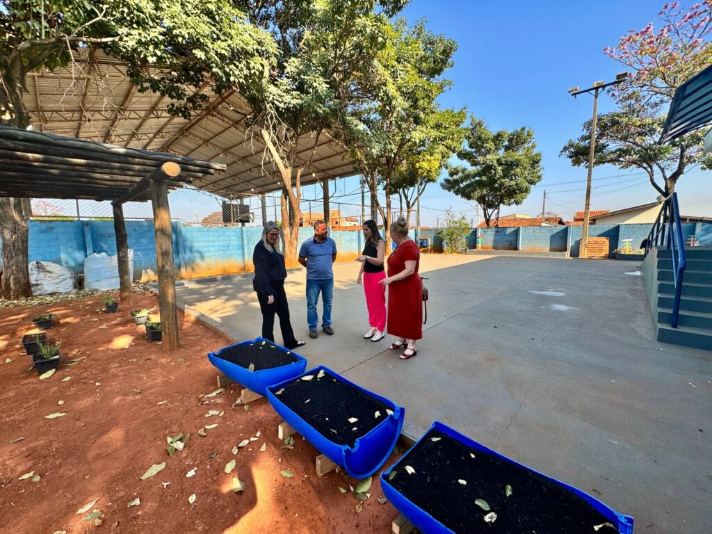 A imagem mostra uma área externa onde quatro pessoas estão reunidas e conversando próximas a jardineiras retangulares azuis, preenchidas com terra e algumas pequenas plantas. O ambiente parece pertencer a uma escola ou centro comunitário, com construções ao redor, escadas que levam a um prédio e um chão que mistura concreto com terra vermelha. Árvores oferecem sombra ao local, e ao fundo há uma cobertura metálica sustentada por pilares, além de um muro azul que delimita o espaço.
