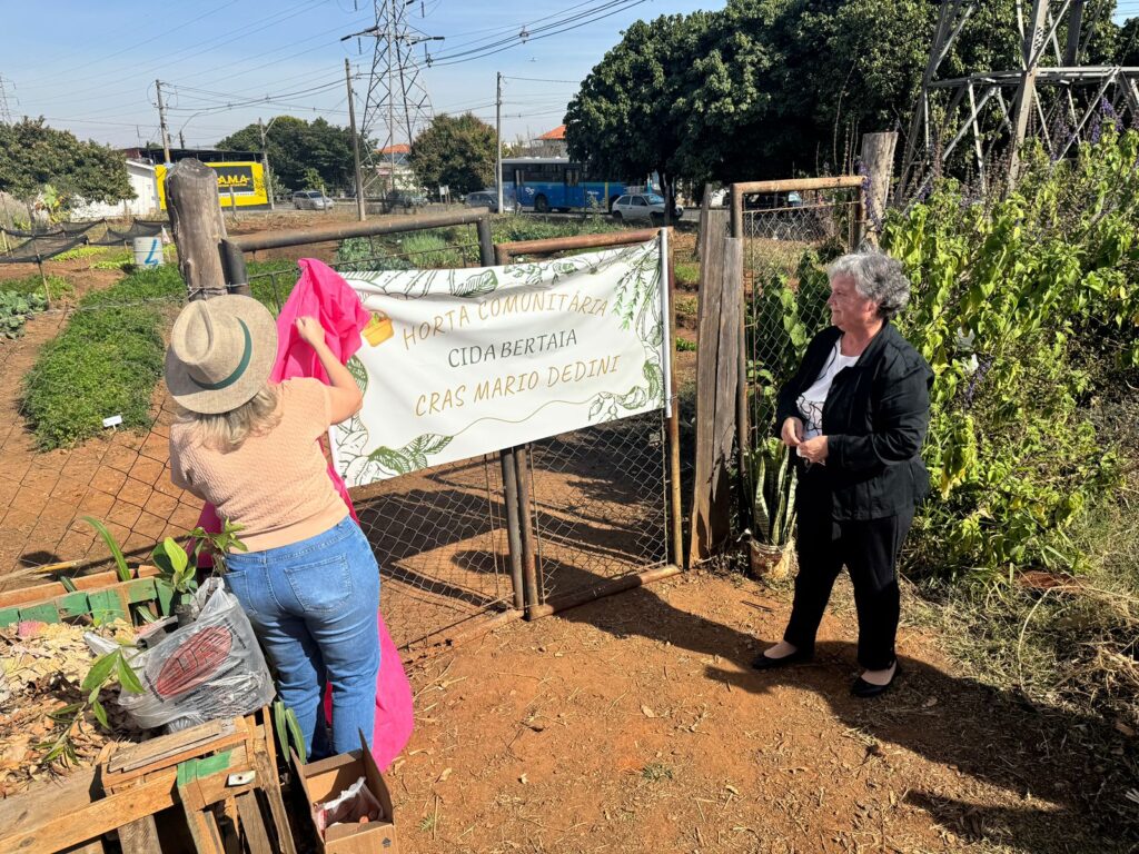 A imagem mostra duas pessoas diante da entrada da Horta Comunitária Cida Bertatia – CRAS Mario Dedini, em um ambiente urbano com ônibus e prédios ao fundo. Uma delas segura um pano rosa, como se estivesse prestes a revelar uma placa, enquanto a outra observa.