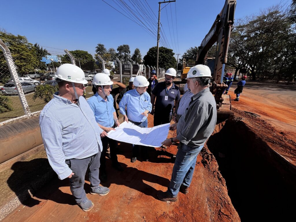 Um grupo de homens, todos usando capacetes brancos, está reunido em torno de um projeto. Eles parecem estar discutindo o projeto, pois um deles está segurando o projeto aberto. O cenário é um local de construção, com um grande escavadeira Volvo em segundo plano. Há um buraco ou vala na frente do grupo, com terra vermelha ao redor. A luz do sol está forte, criando sombras nítidas. No lado esquerdo da imagem, há uma cerca de metal com uma área de estacionamento e alguns carros atrás dela. Há também algumas árvores e um edifício ao fundo. O céu está claro e azul. Os homens estão vestidos com roupas casuais, incluindo camisas e calças jeans. Alguns deles estão usando camisas de mangas compridas, enquanto outros estão usando camisas de mangas curtas. Todos estão usando capacetes de segurança brancos, que são um requisito para trabalhar em um local de construção. A atmosfera geral da imagem é de profissionalismo e foco. Os homens parecem estar engajados em sua discussão e dedicados ao seu trabalho.