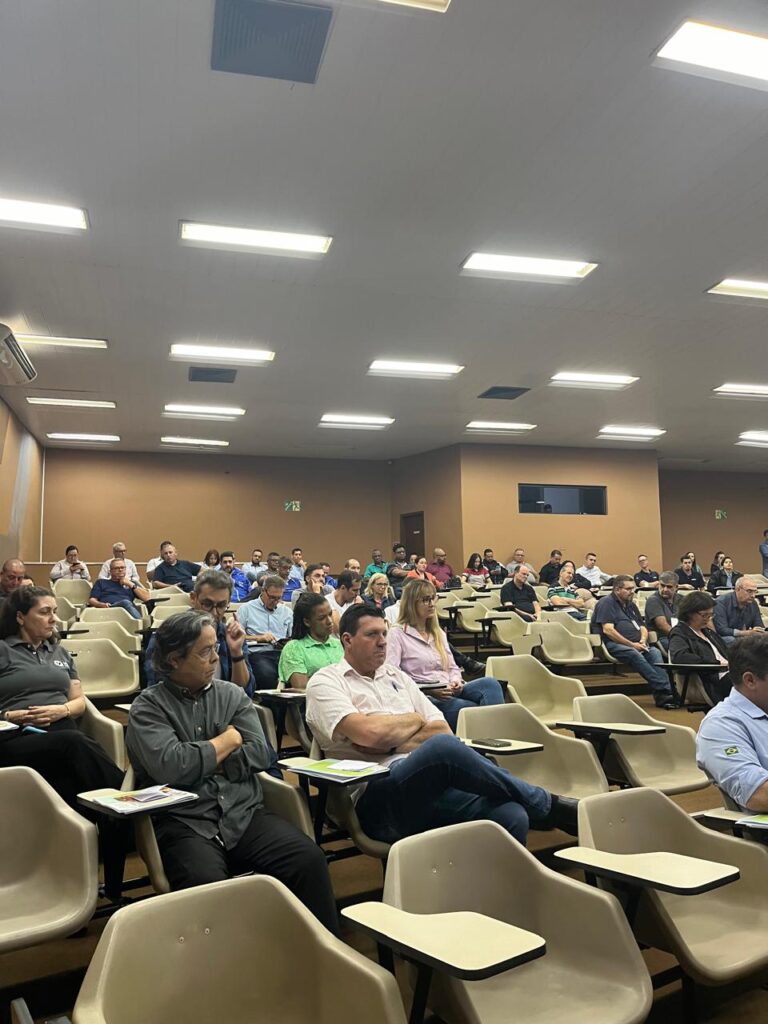 A imagem mostra uma sala de aula ou auditório com várias pessoas sentadas em cadeiras, prestando atenção em algo fora do enquadramento. A maioria das pessoas parece ser adulta e está vestida de forma casual ou semi-formal. O ambiente é iluminado por lâmpadas fluorescentes no teto, e as paredes são de cor bege. Algumas pessoas estão com cadernos ou papéis nas mesas à frente das cadeiras. A perspectiva da foto é de alguém sentado na plateia, olhando para a frente.
