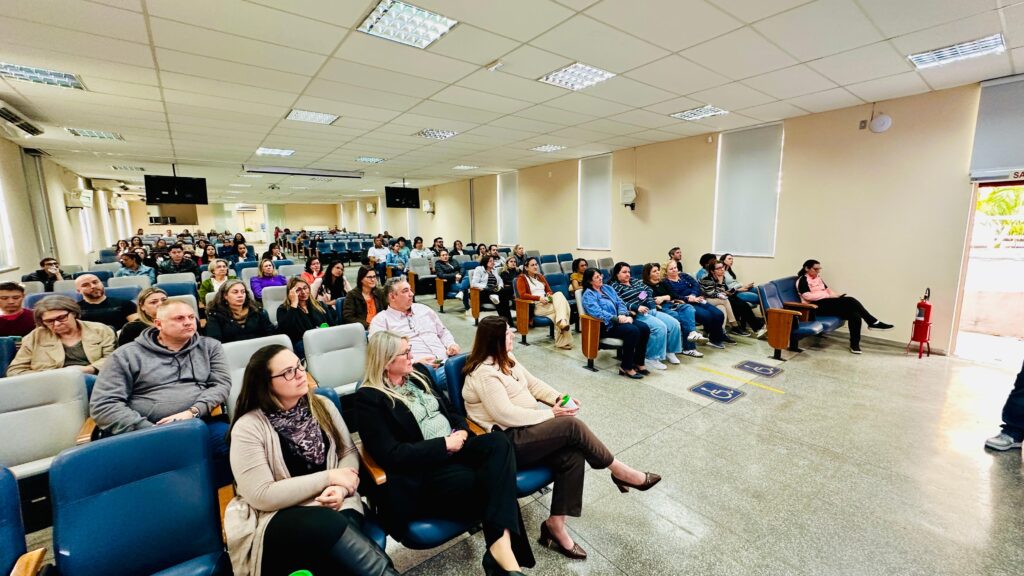 A imagem mostra um auditório cheio, com diversas pessoas sentadas em fileiras, todas voltadas para a frente, onde provavelmente ocorre uma apresentação ou palestra. O ambiente é bem iluminado por luzes fluorescentes no teto, e há duas grandes telas suspensas, reforçando o caráter informativo do evento. As paredes são bege, com janelas parcialmente cobertas por persianas, e o piso é revestido com azulejos claros. Há sinalização de acessibilidade e um extintor de incêndio visível próximo à entrada lateral.