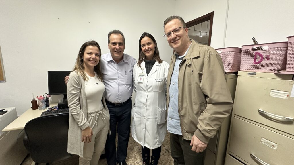 Na imagem, quatro pessoas posam juntas em um escritório. A pessoa mais à esquerda é uma mulher com cabelos castanhos, usando um conjunto de moletom cinza claro e um cardigã por cima. Ela está sorrindo para a câmera. Ao lado dela, um homem de camisa clara e calça escura também sorri. No centro da imagem, há uma mulher vestindo um jaleco branco de enfermeira. Seu nome, "Cátia Dusali", e sua profissão, "Enfermeira", estão escritos em uma etiqueta em seu jaleco. Ela tem cabelos escuros e sorri para a câmera. A pessoa mais à direita é um homem de óculos, usando uma jaqueta bege sobre uma camisa azul e calça escura. Ele também está sorrindo. Ao fundo, à esquerda, há uma mesa de escritório com um monitor de computador, um teclado e alguns objetos em cima. À direita, há arquivos e cestas cor-de-rosa empilhadas.