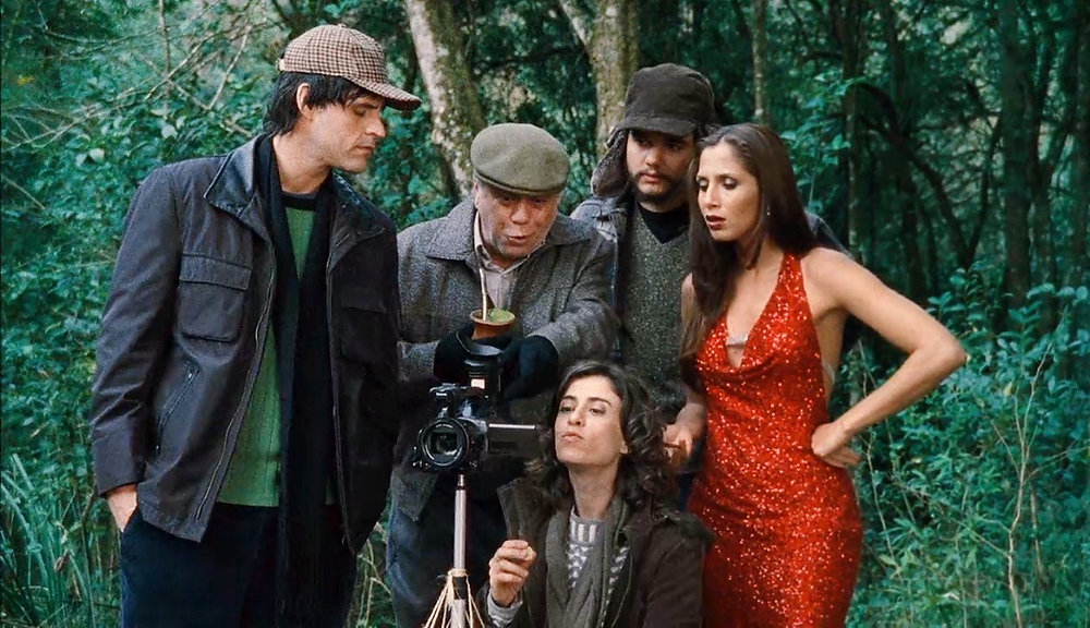 Grupo de cinco pessoas em uma floresta observa uma câmera; ao centro, uma mulher sentada mexe no equipamento, enquanto os outros, incluindo uma mulher de vestido vermelho brilhante, acompanham atentos.